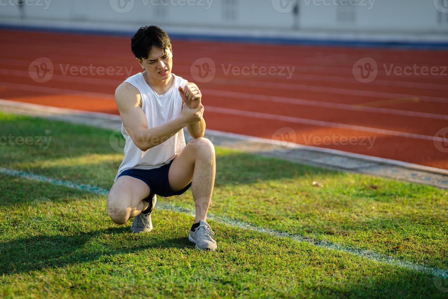 White tank top asian man is holding his wrist while crouching on grass field. Pain or injury. photo