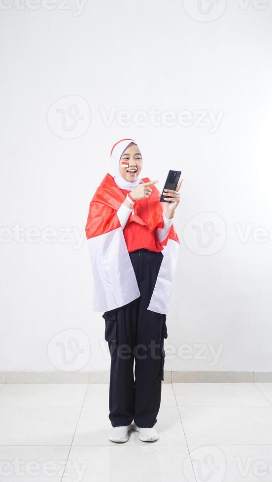 Indonesia independence day celebration with young adult Asian girl wearing red and white Indonesia flag color attribute holding and pointing at smartphone isolated on white background photo