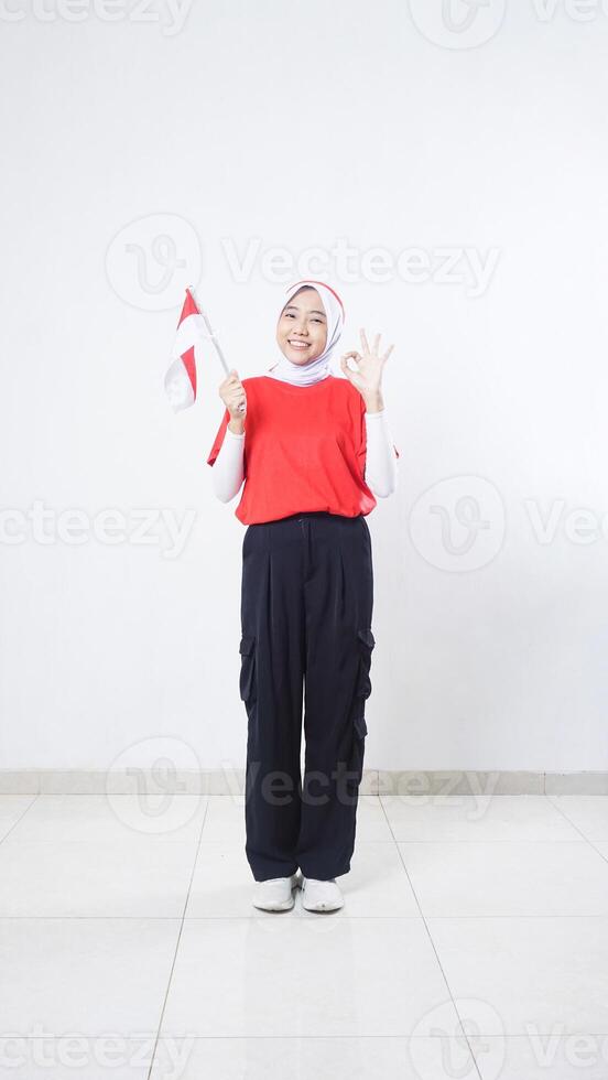 Indonesia independence day celebration with Asian girl wearing red and white outfit holding mini Indonesia flag decoration and showing OK hand gesture sign isolated on white background photo