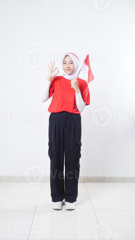 Indonesia independence day celebration with smiling Asian girl wearing red and white outfit holding mini Indonesia flag decoration and showing OK hand gesture sign isolated on white background photo