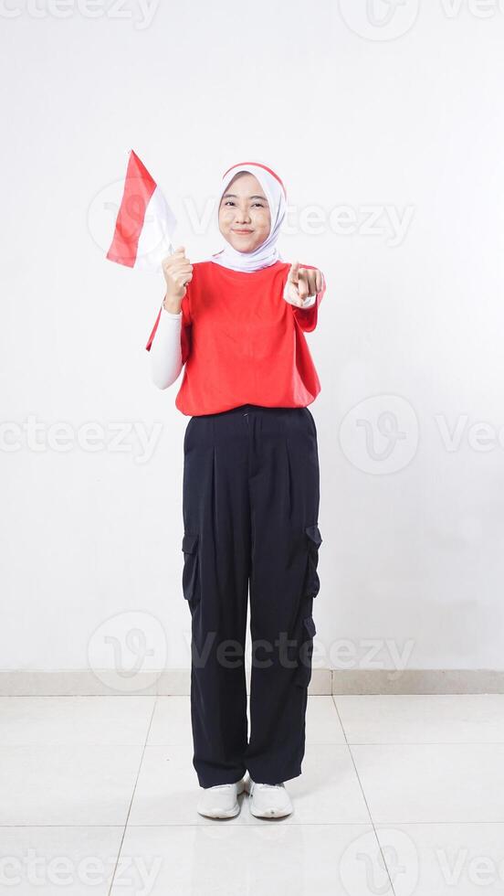 Indonesia independence day celebration with Asian girl wearing red and white outfit holding mini Indonesia flag decoration and pointing at camera with her index finger isolated on white background photo