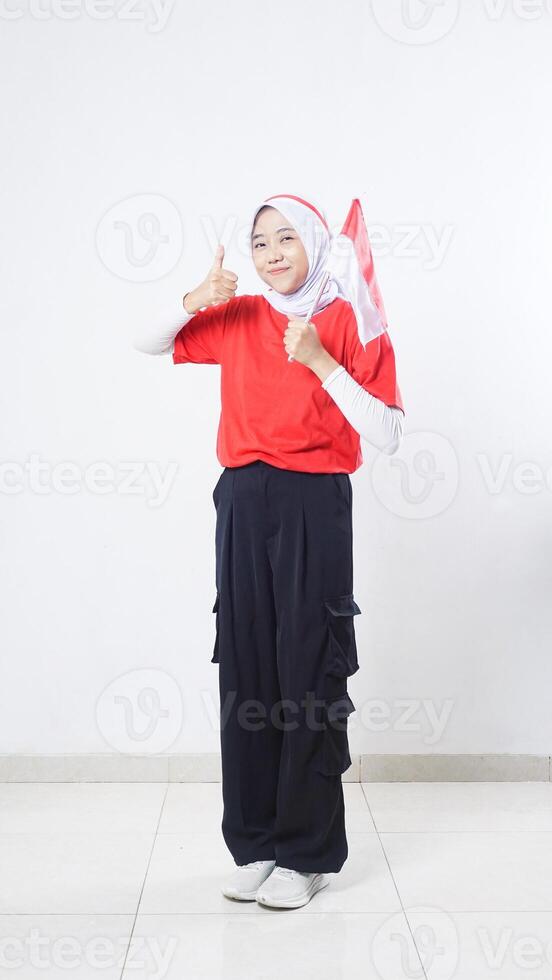 Indonesia independence day celebration with beautiful Asian girl wearing red and white outfit holding mini Indonesia flag decoration and showing her thumb up isolated on white background photo