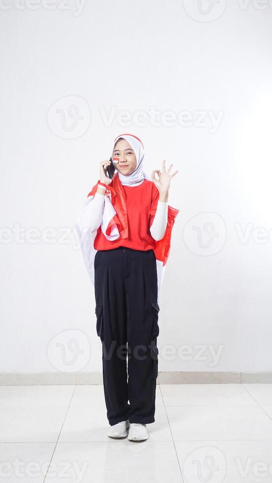 Indonesia independence day celebration with Asian girl wearing red and white Indonesia flag color attribute talking on the phone call and showing OK sign with hand gesture isolated on white background photo