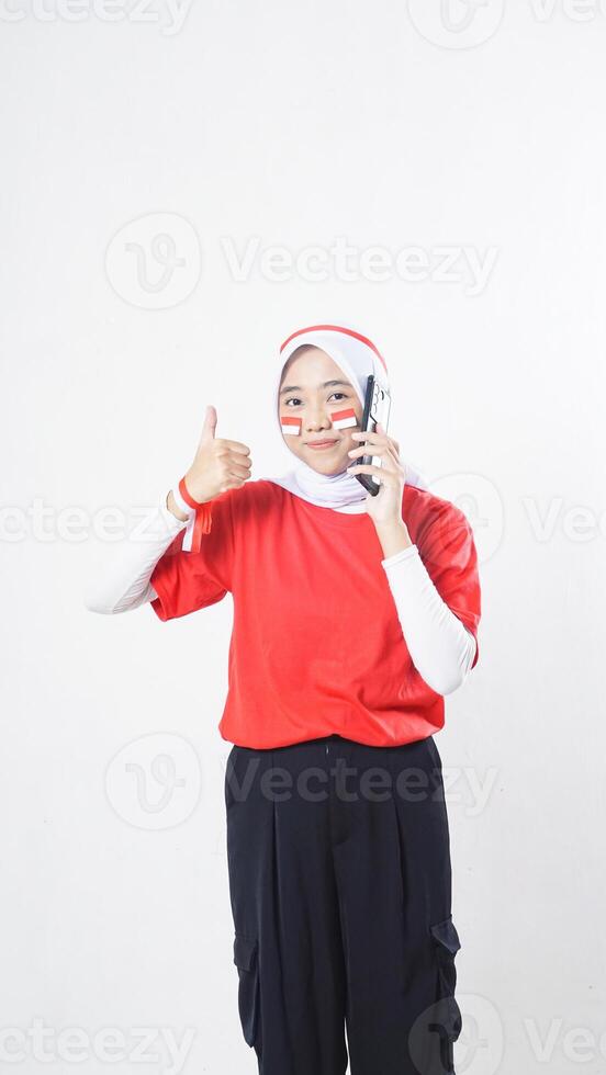 Indonesia independence day celebration with smiling Asian girl wearing red and white Indonesia flag color attribute talking on phone call while raising her thumb up isolated on white background photo