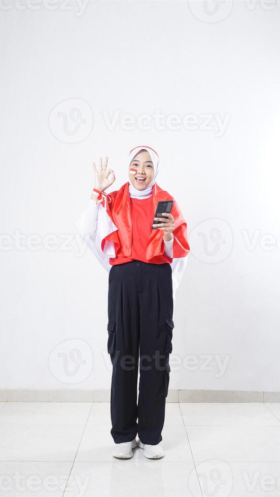 Indonesia independence day celebration with happy Asian girl wearing red and white Indonesia flag color attribute holding smartphone and showing OK sign with hand gesture isolated on white background photo