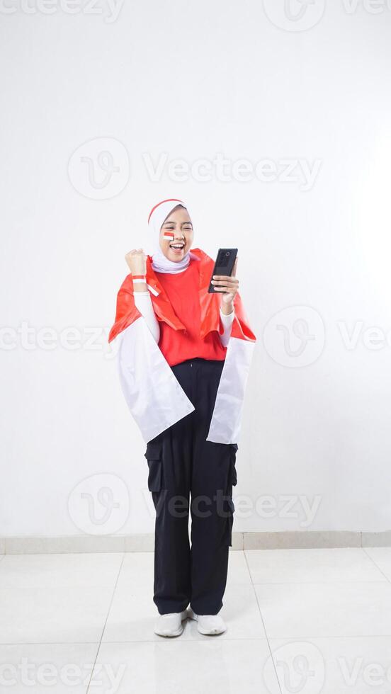 Indonesia independence day celebration with excited Asian girl wearing red and white Indonesia flag color attribute holding smartphone and raising a hand clenched isolated on white background photo