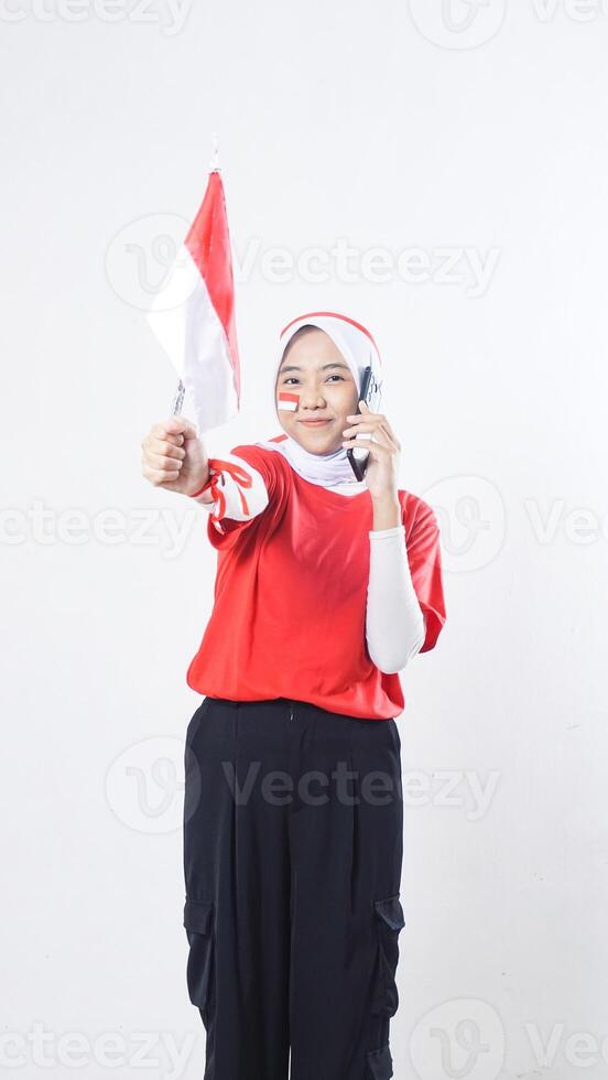 Indonesia independence day celebration with Asian girl wearing red and white Indonesia flag color attribute talking on phone call while showing Indonesia flag decoration isolated on white background photo