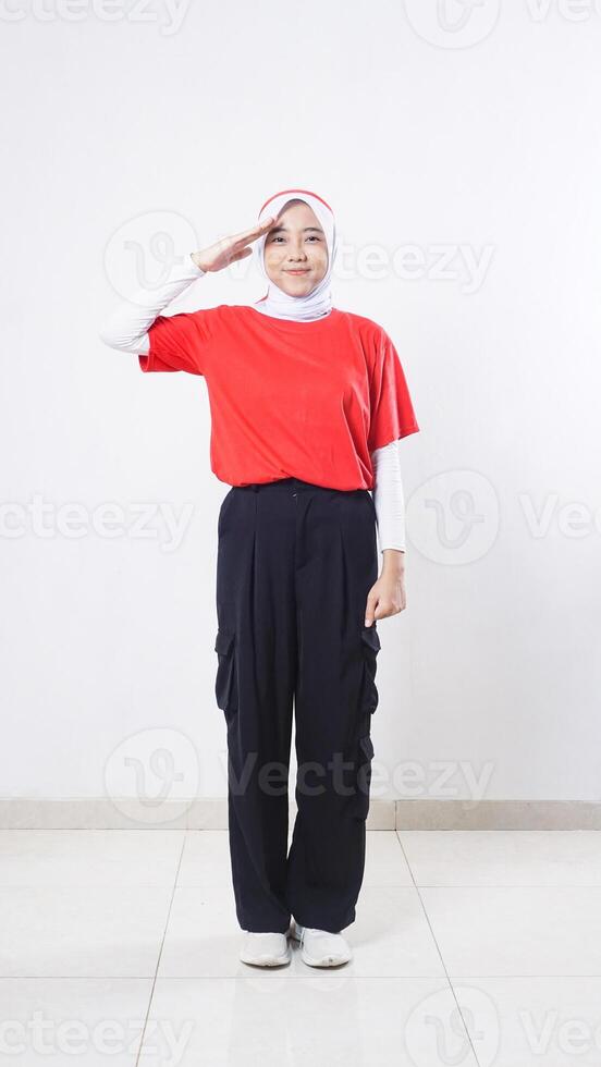 Asian girl celebrate Indonesia independence day with red and white outfit doing salute pose with right hand raised to the eyebrow isolated on white background photo