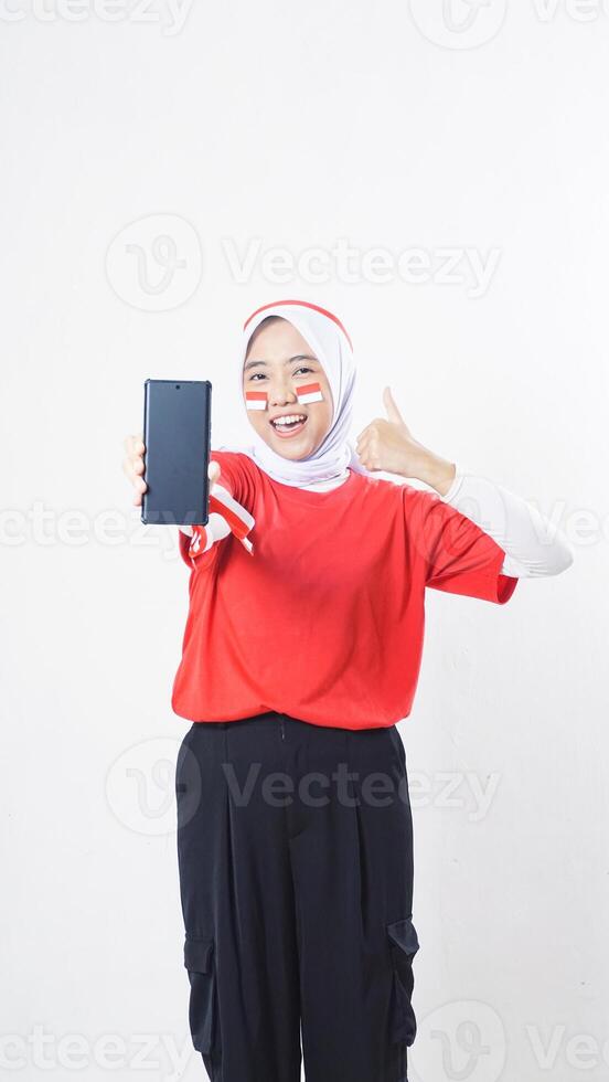 Indonesia independence day celebration with excited Asian girl wearing red and white Indonesia flag color attribute showing smartphone blank screen and her thumb up isolated on white background photo