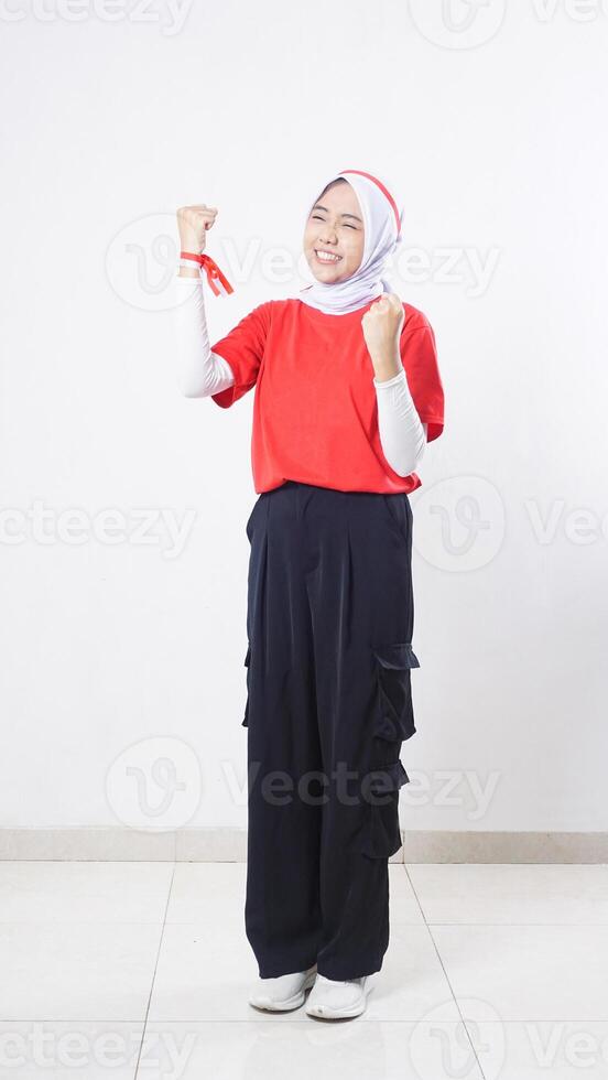 Excited Asian girl celebrate Indonesia independence day with red and white outfit showing winning gesture with raising both her clenched fists isolated on white background photo