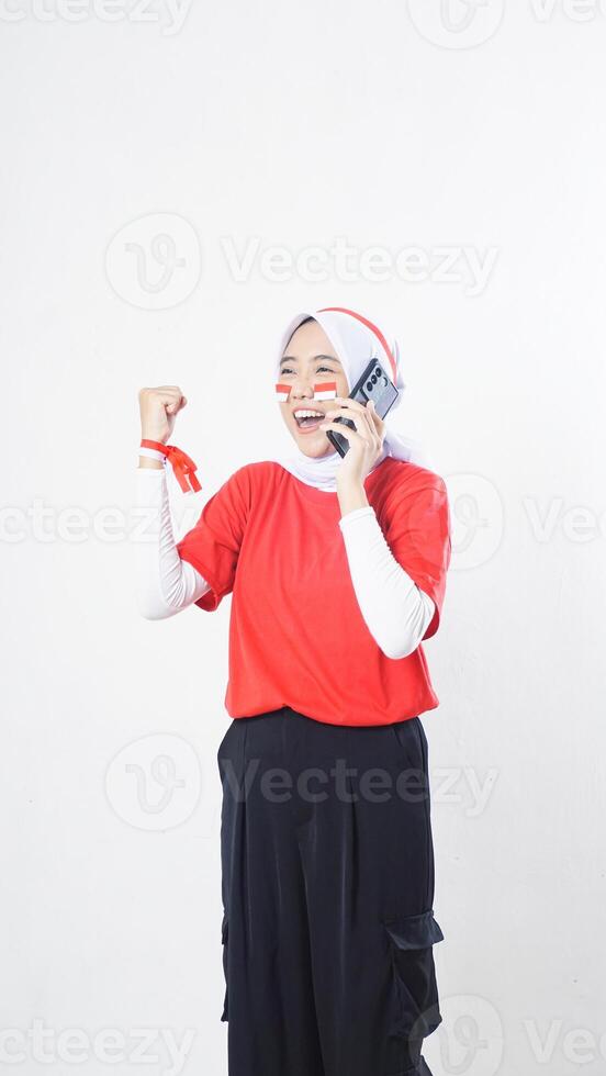 Indonesia independence day celebration with excited Asian girl wearing red and white Indonesia flag color attribute talking on phone call while raising her hand clenched isolated on white background photo
