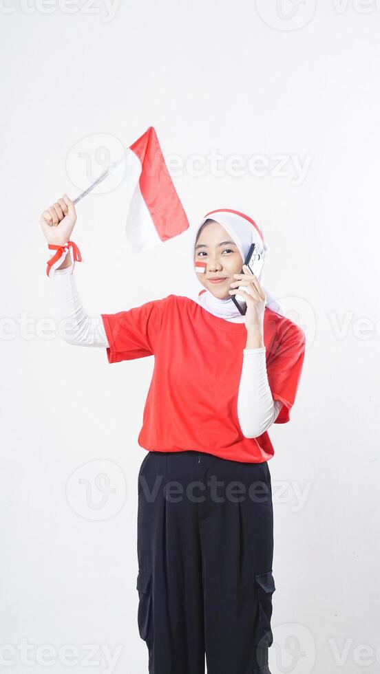 Indonesia independence day celebration with Asian girl wearing red and white Indonesia flag color attribute talking on phone call while raising Indonesia flag decoration isolated on white background photo