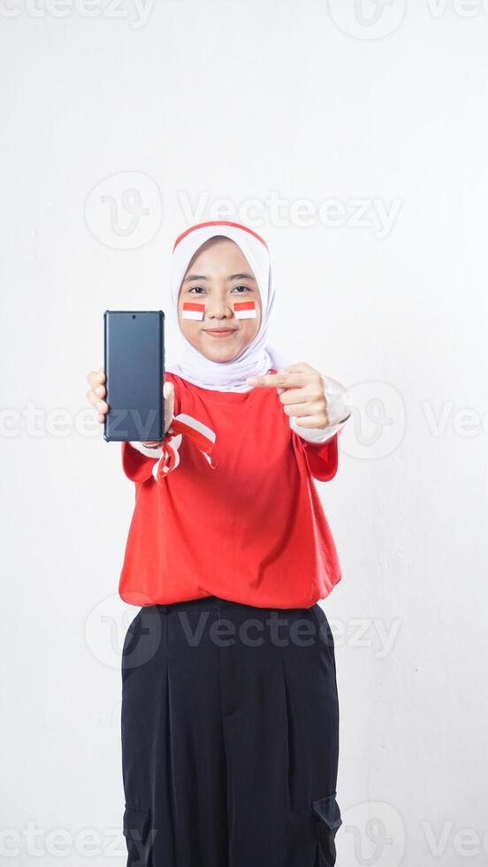 Indonesia independence day celebration with Asian girl wearing red and white Indonesia flag color attribute showing and pointing at smartphone with blank screen isolated on white background photo