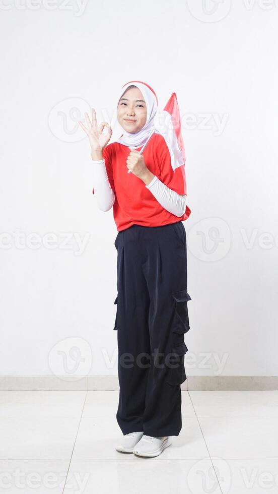 Indonesia independence day celebration with beautiful Asian girl wearing red and white outfit holding mini Indonesia flag decoration and showing OK hand gesture sign isolated on white background photo