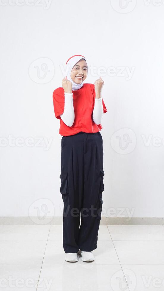 Asian girl celebrate Indonesia independence day with red and white outfit doing winning gesture with clenching both her fists isolated on white background photo