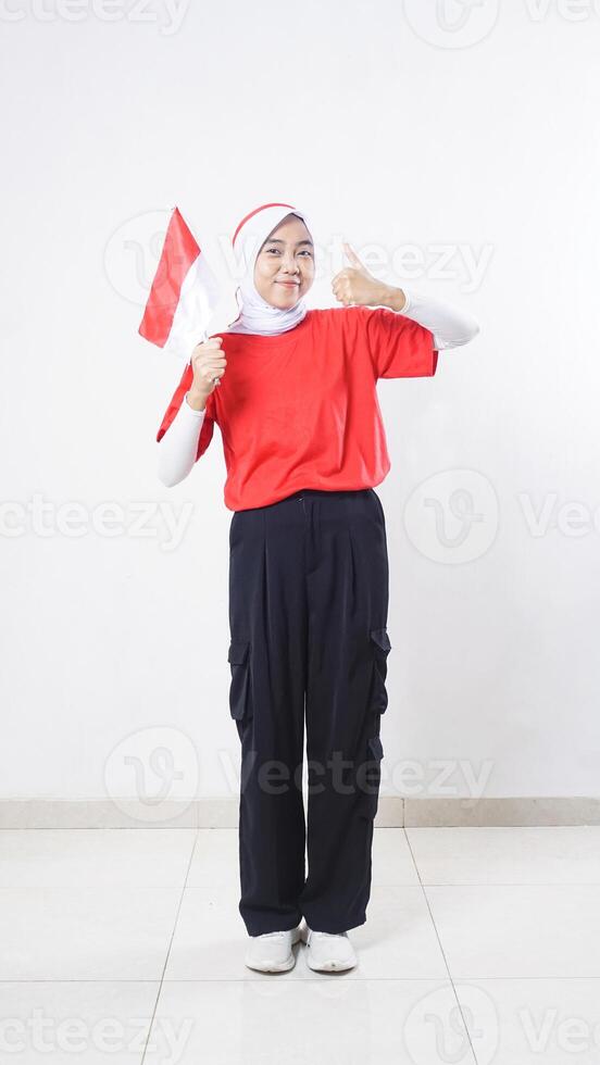 Indonesia independence day celebration concept with beautiful Asian girl wearing red and white outfit holding mini Indonesia flag decoration and showing her thumb up isolated on white background photo