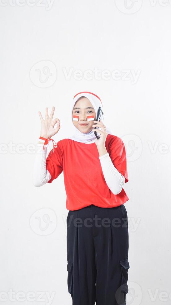 Indonesia independence day celebration with smiling Asian girl wearing red and white Indonesia flag color attribute talking on phone call while showing OK hand gesture isolated on white background photo