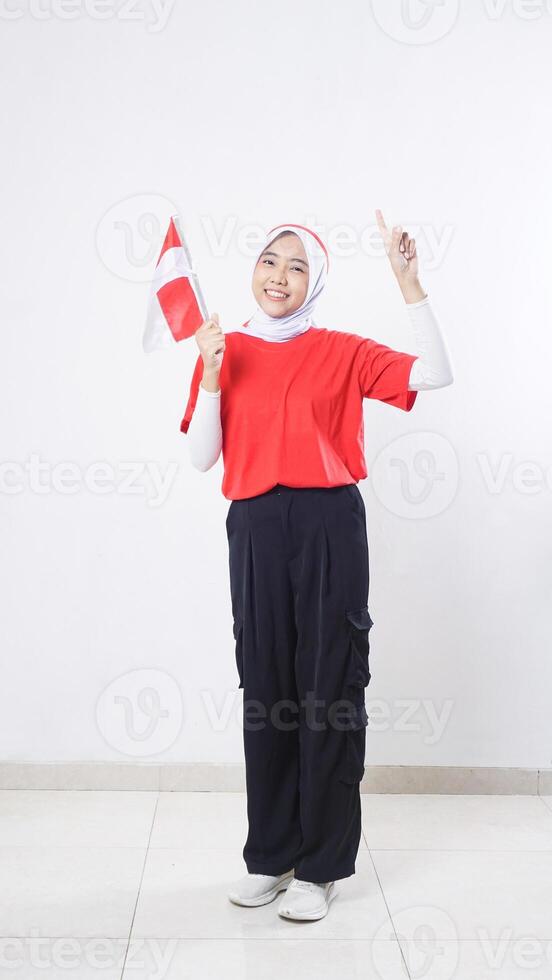 Indonesia independence day celebration with beautiful Asian girl wearing red and white outfit holding mini Indonesia flag decoration and raising her index finger up isolated on white background photo