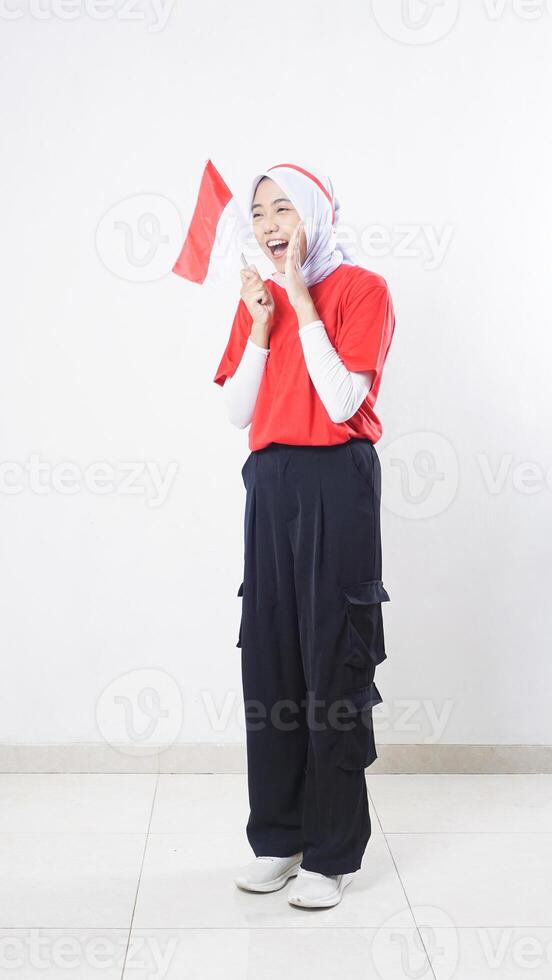 Indonesia independence day celebration with excited Asian girl wearing red and white outfit holding mini Indonesia flag decoration and doing shout pose isolated on white background photo