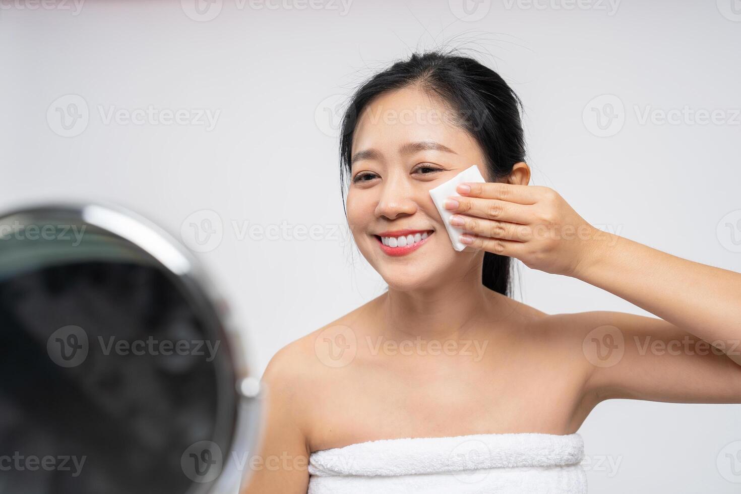 Happy asian woman looking at mirror and using cotton pad clean make up on her face photo