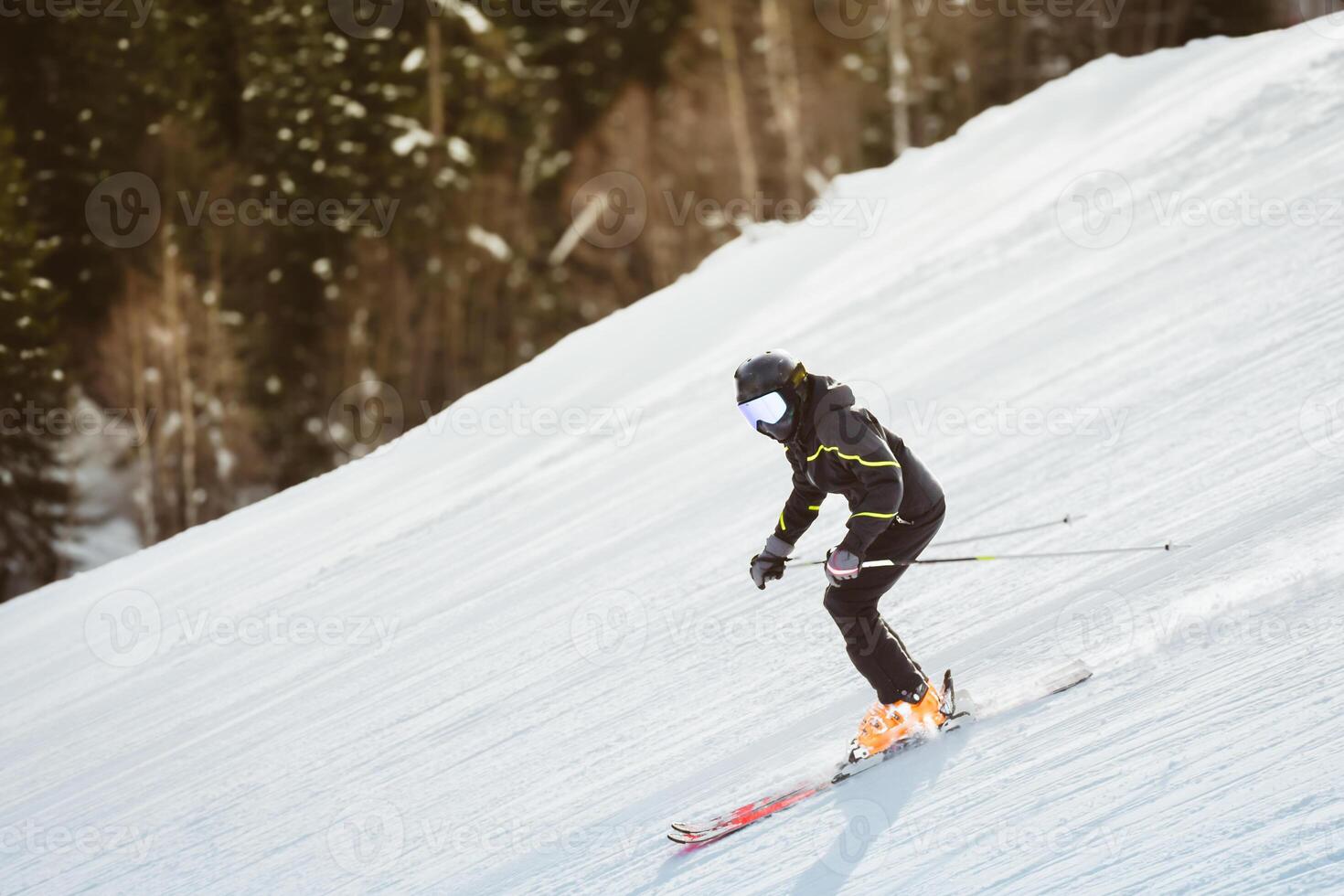 Close up freeze motion skier wearing helmet goggles carving on downhill looking at camera snowy slope pine forest background fast winter sport action concept. Unrecognizable intentional blur photo