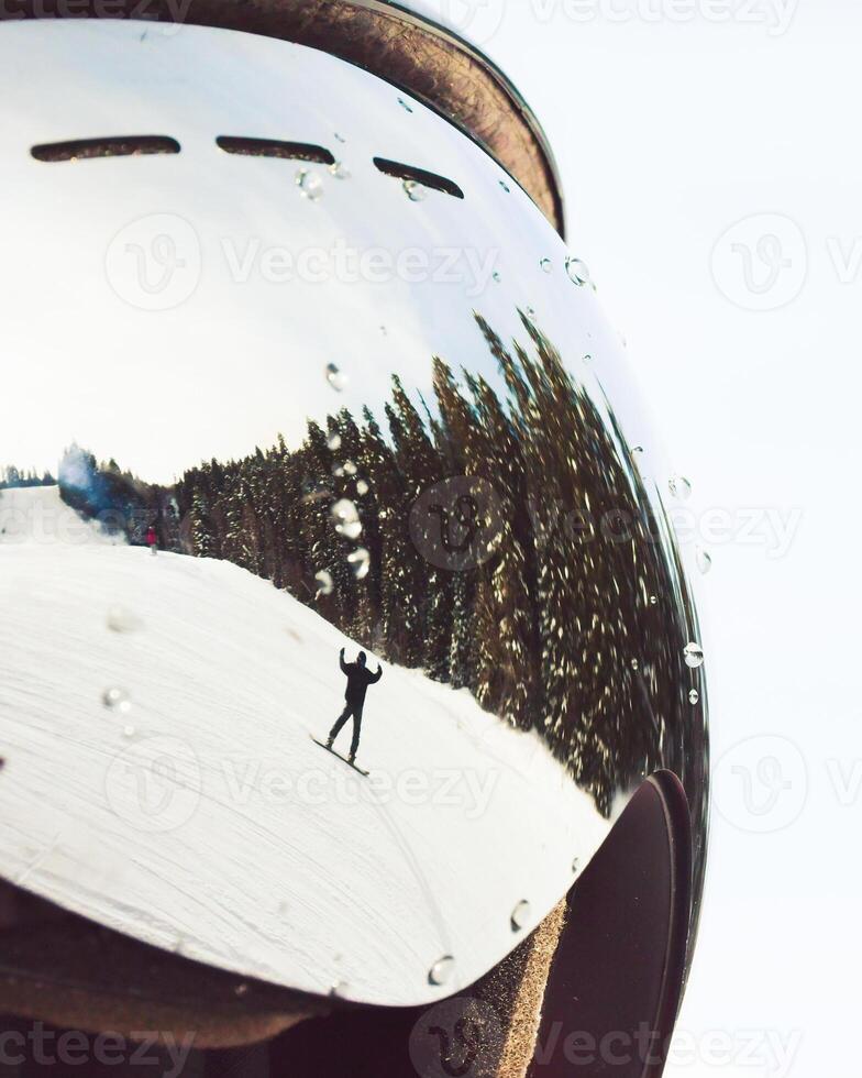 Snowboarder carving snowy slope reflected in ski goggles with water drops, pine forest under clear winter sky, concept extreme sport, safety, perspective. photo