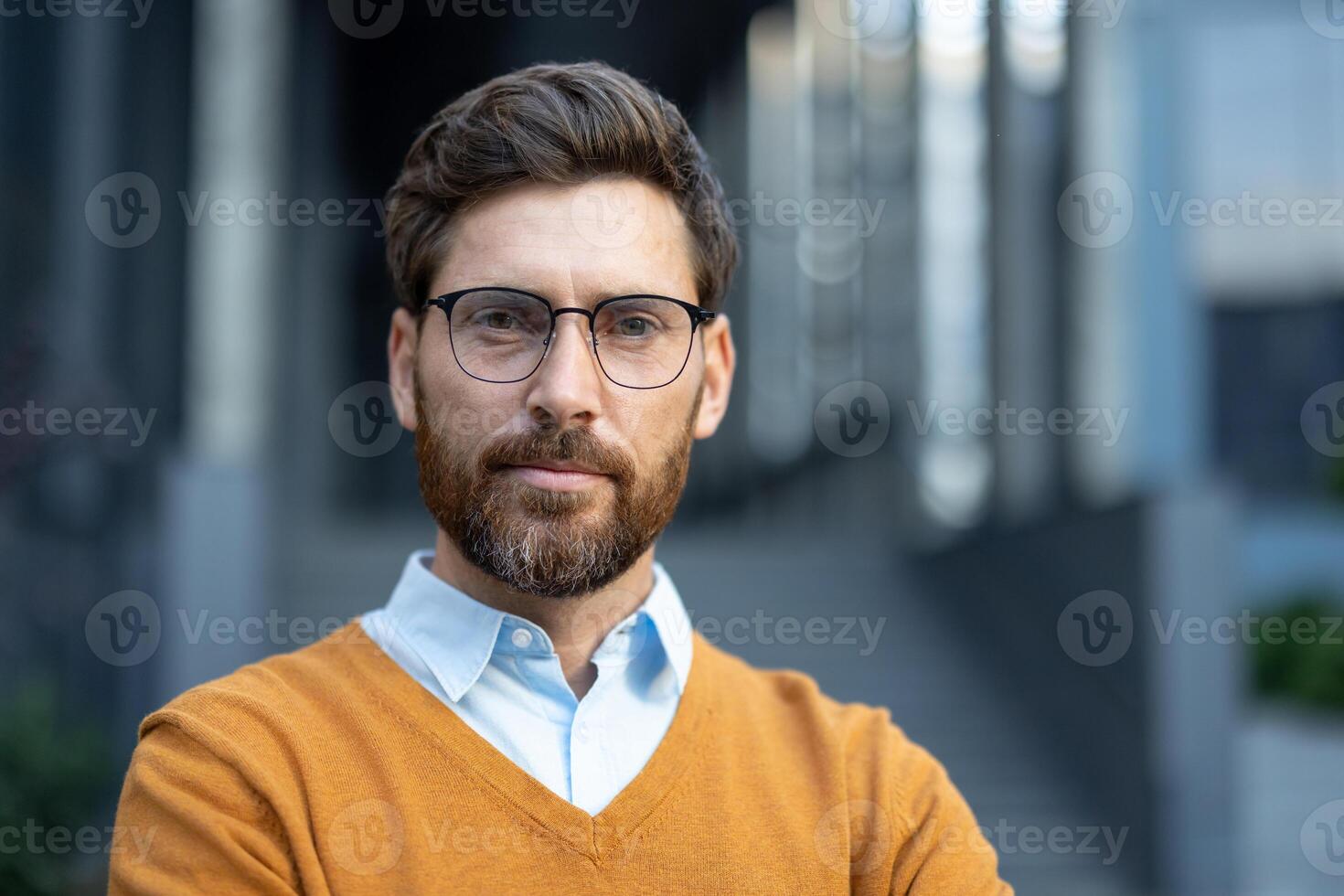 A portrait of a man with glasses and a beard, wearing a yellow sweater and a light blue shirt, taken outdoors. photo