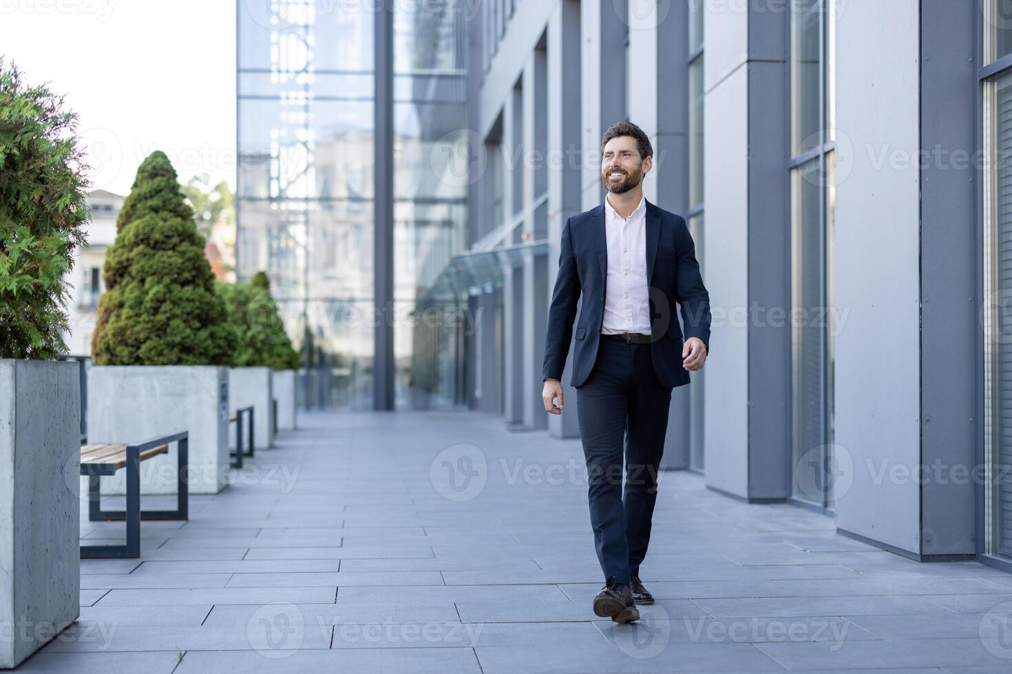 A smiling businessman in a suit walks confidently on an outdoor office walkway, with modern architecture in the background. photo