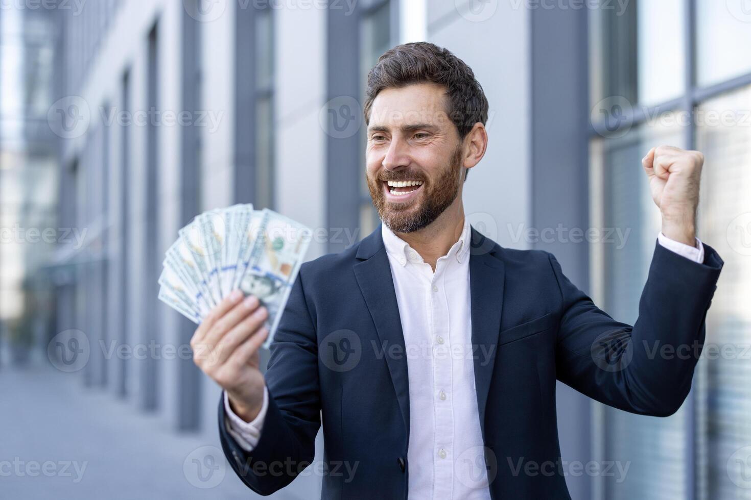 A successful businessman celebrates a financial achievement, holding cash with a triumphant expression against an office building backdrop. photo