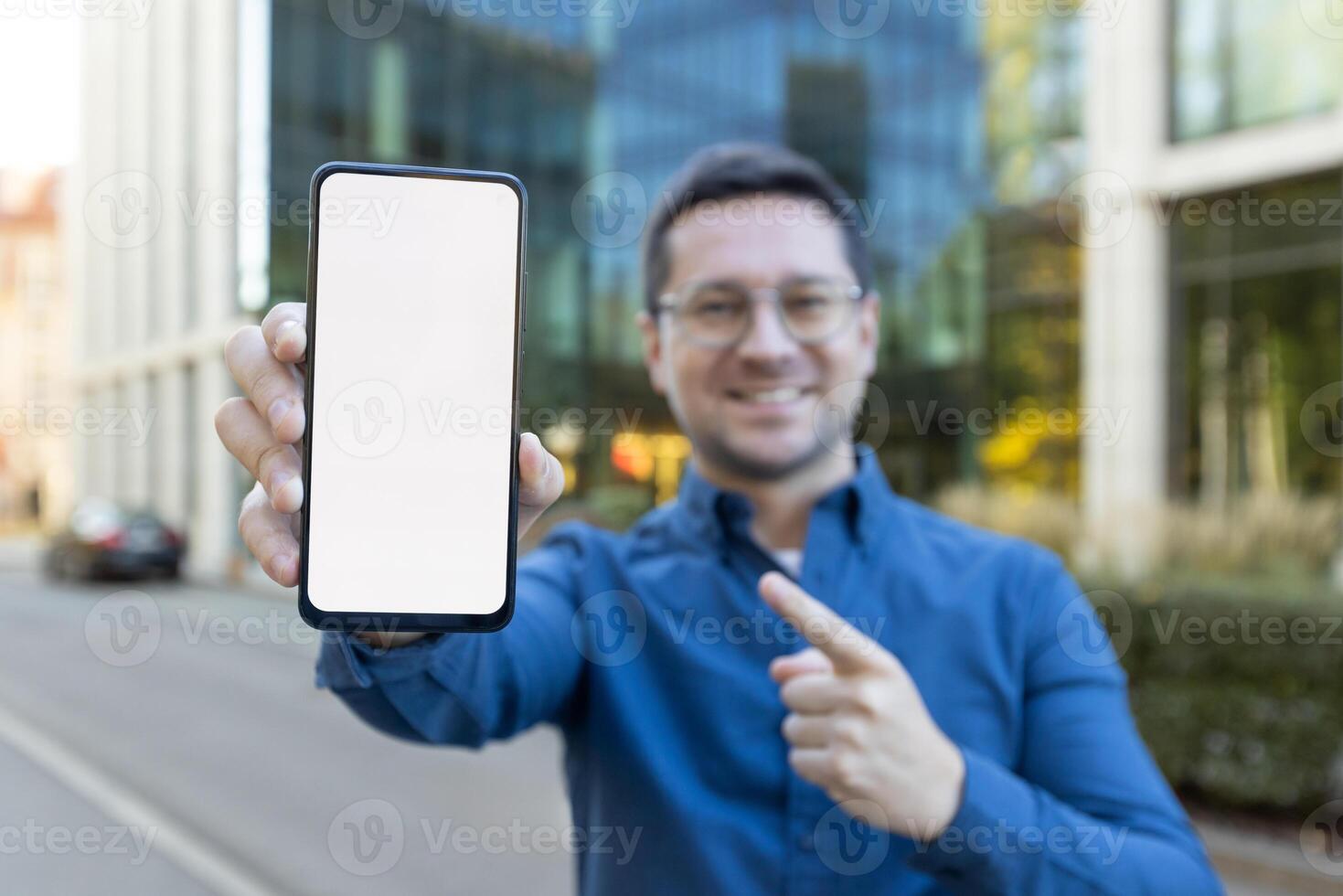 A man holds a phone with a blank screen, pointing at it in a business district. photo