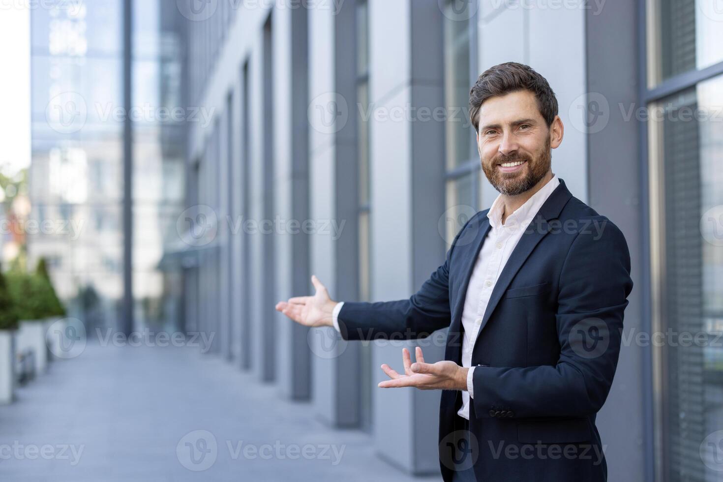 A businessman in a suit gestures with both hands, welcoming or presenting something in front of a modern building. photo