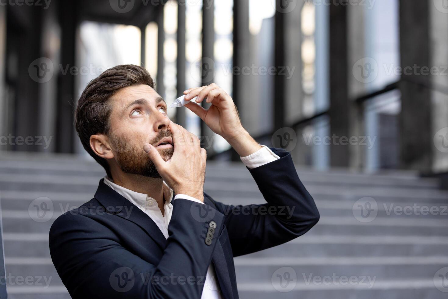 A businessman applying eye drops outdoors. He is wearing a suit and looking up with his mouth open. photo