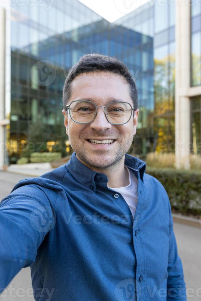 A man wearing glasses takes a selfie in front of a modern glass building, smiling towards the camera. photo