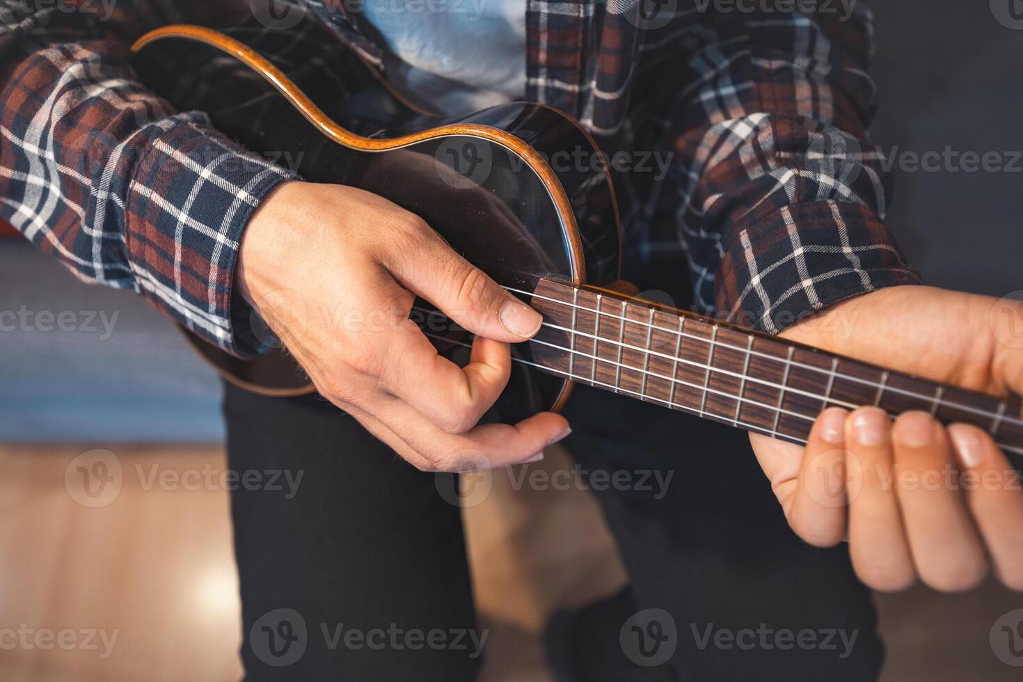 A youth in a plaid shirt strums a ukulele on a grey couch with red pillow photo