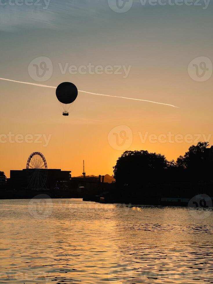 Silhouette of hot air balloon flying over river at sunset with ferris wheel and city skyline in background, creating serene and magical atmosphere. Hot air balloon, ferris wheel and urban landscape photo