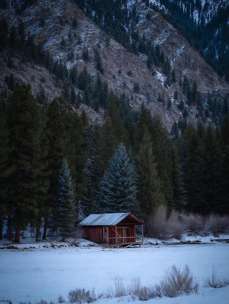 Secluded Cabin in a Snowy Mountain Landscape. A rustic red cabin sits peacefully amidst a snow-covered field, surrounded by towering evergreen trees and a dramatic mountain backdrop. photo