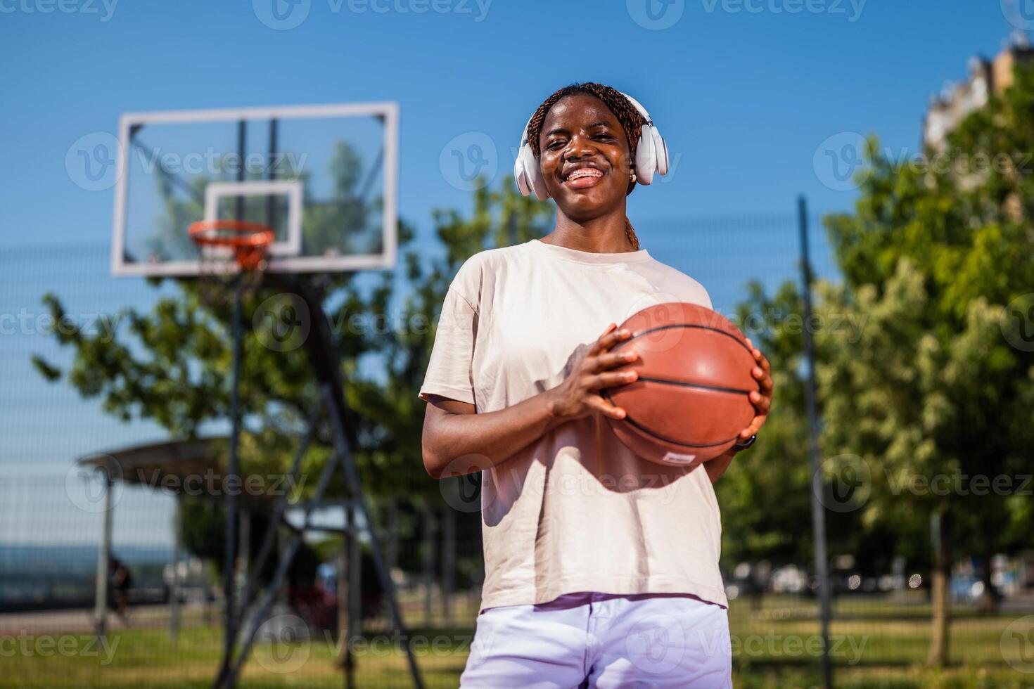 Young black female athlete takes a break during practice, smiling and enjoying music through headphones while holding a basketball on a city court. photo