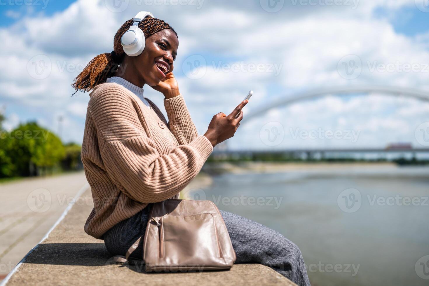 Beautiful African American woman sitting outdoors, casually enjoying music with wireless headphones while texting on her phone, embodying modern city life and relaxation. photo
