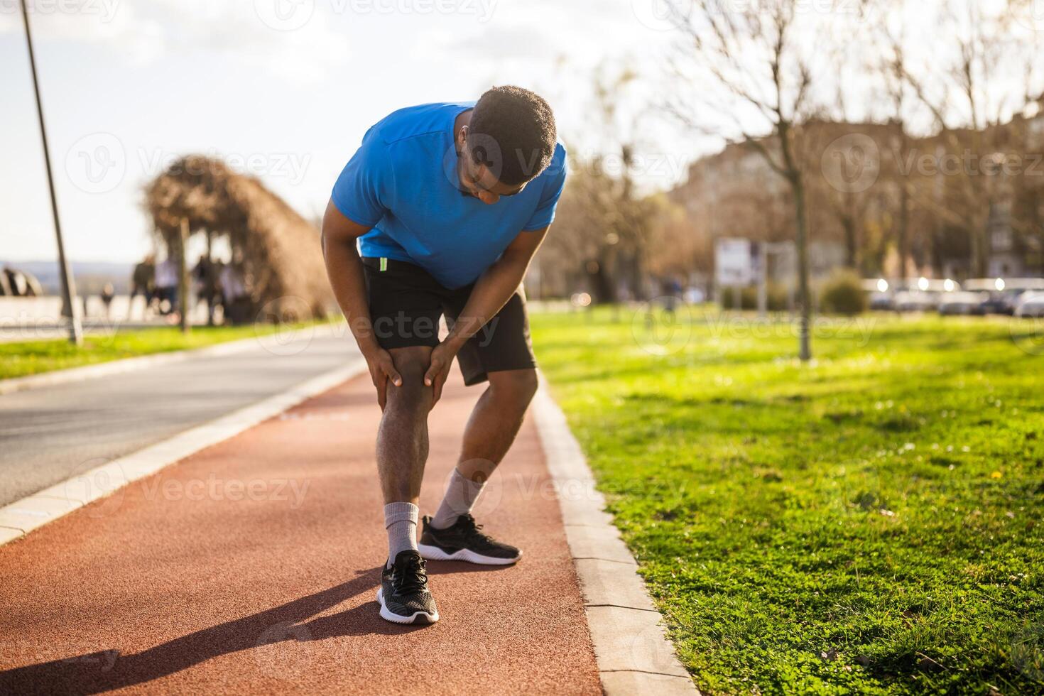 African American male runner experiencing knee pain on a city running track, frowning as he takes a break from exercising. photo