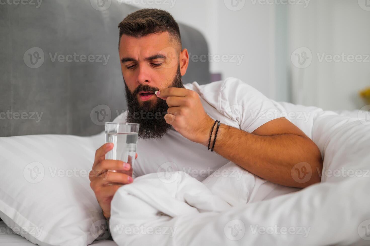 Bearded man holding a pill and a glass of water, preparing to take medication to combat insomnia at bedtime. photo