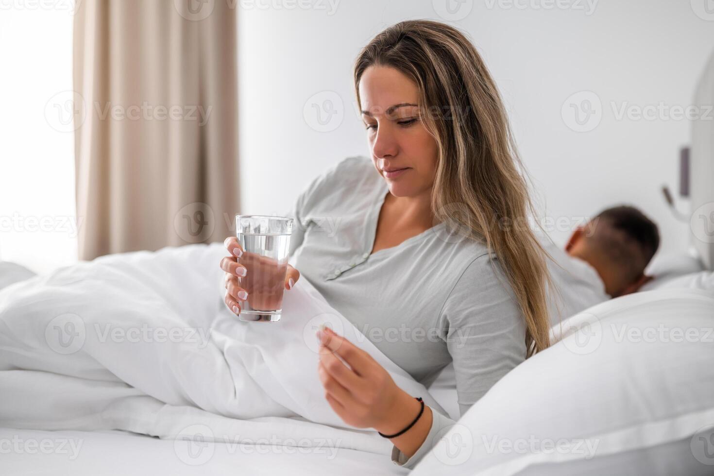 Tired woman in bed, holding pill and glass of water, struggling with insomnia and emotional stress during bedtime. photo