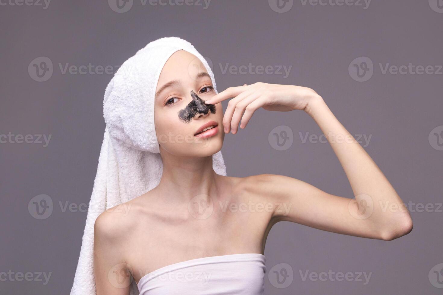 Young caucasian female applying charcoal face mask with towel on head for skincare routine. photo