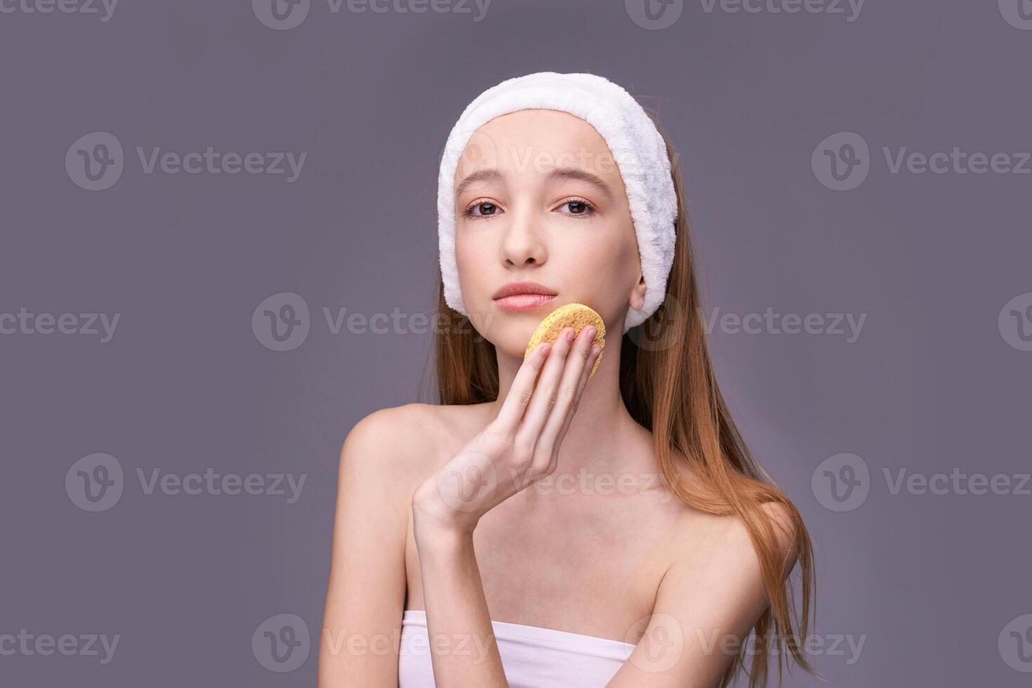 Young caucasian female skincare routine with headband and sponge against gray background. photo