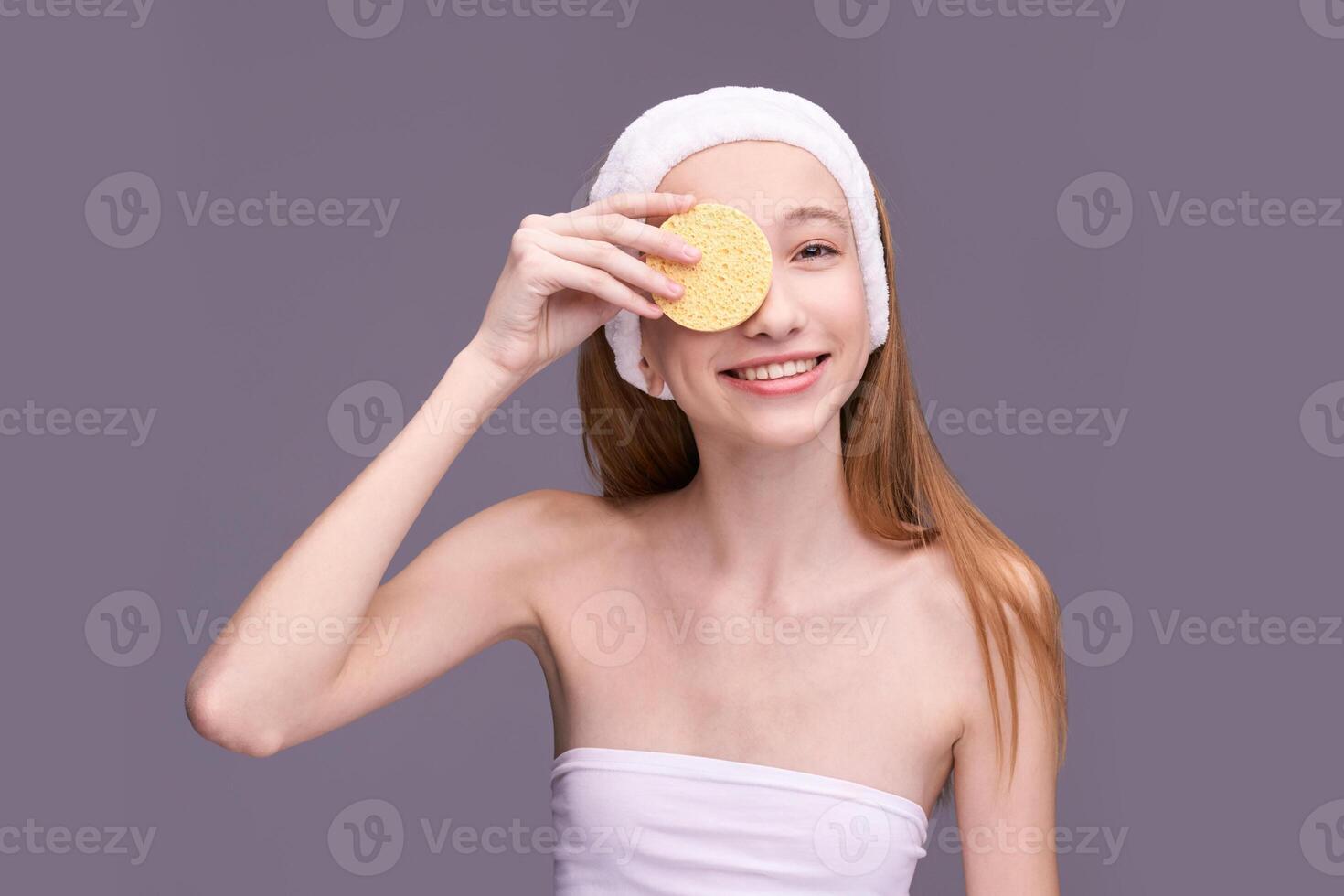 Young caucasian female skincare routine with facial sponge and towel headband on gray background. photo