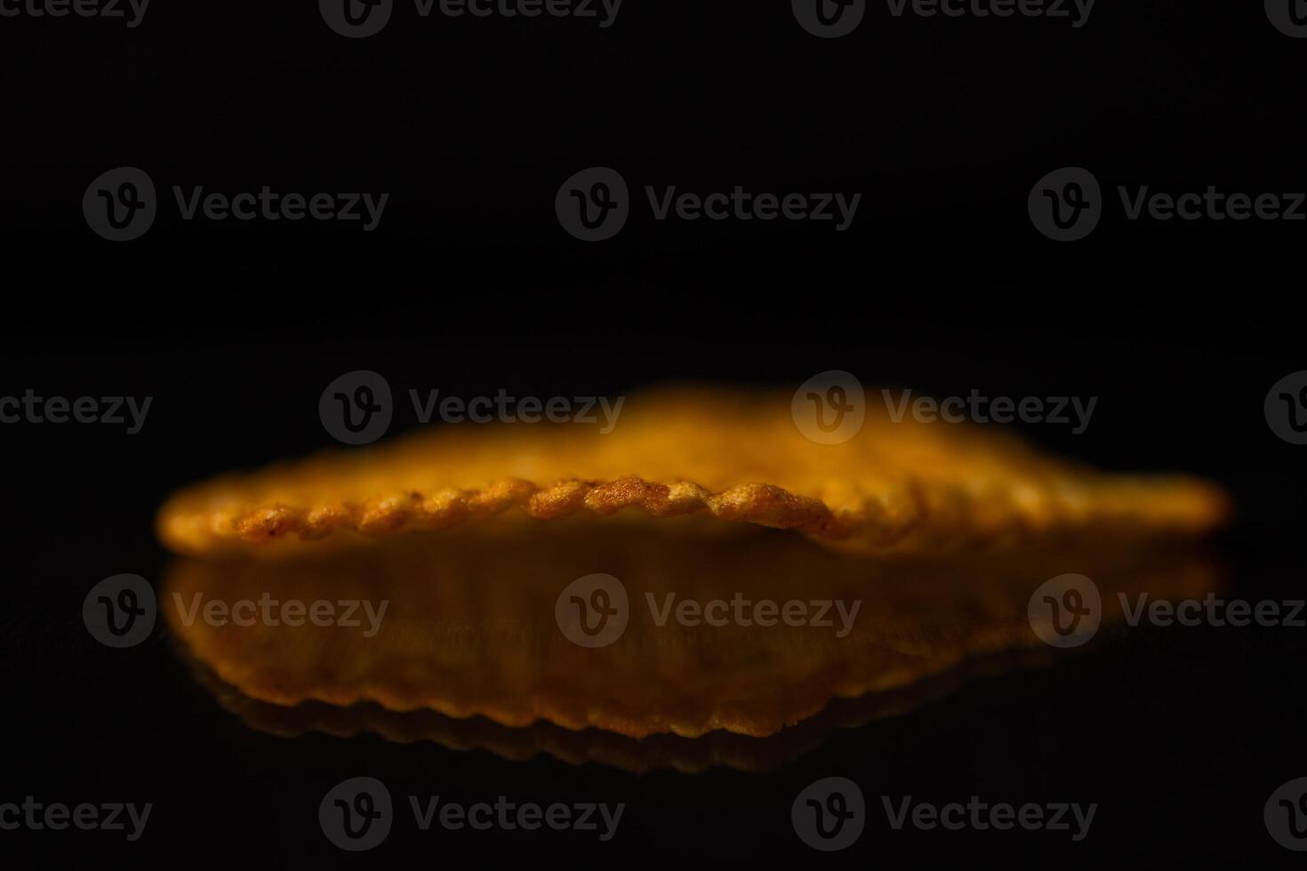 A single golden crispy snack chip displaying a distinct ridged texture, perfectly reflecting on a dark shiny surface, symbolizing indulgent pleasure of unhealthy food photo