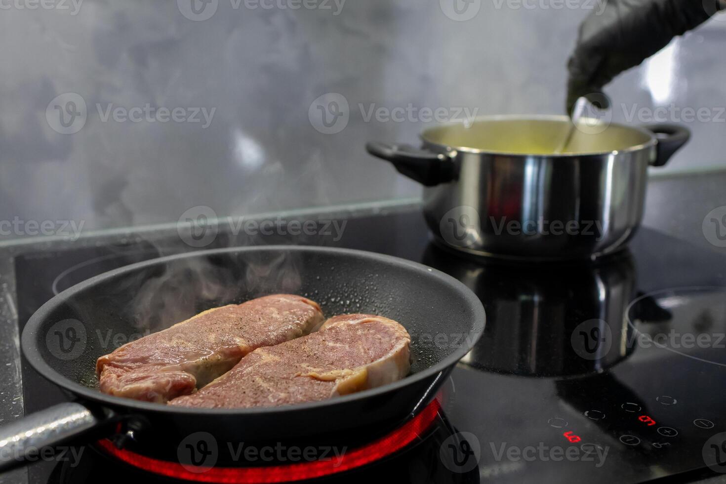 Two raw steaks cooking in a hot pan on an induction hob, with a pot simmering, depicting home cooking, meal preparation, and culinary skill photo