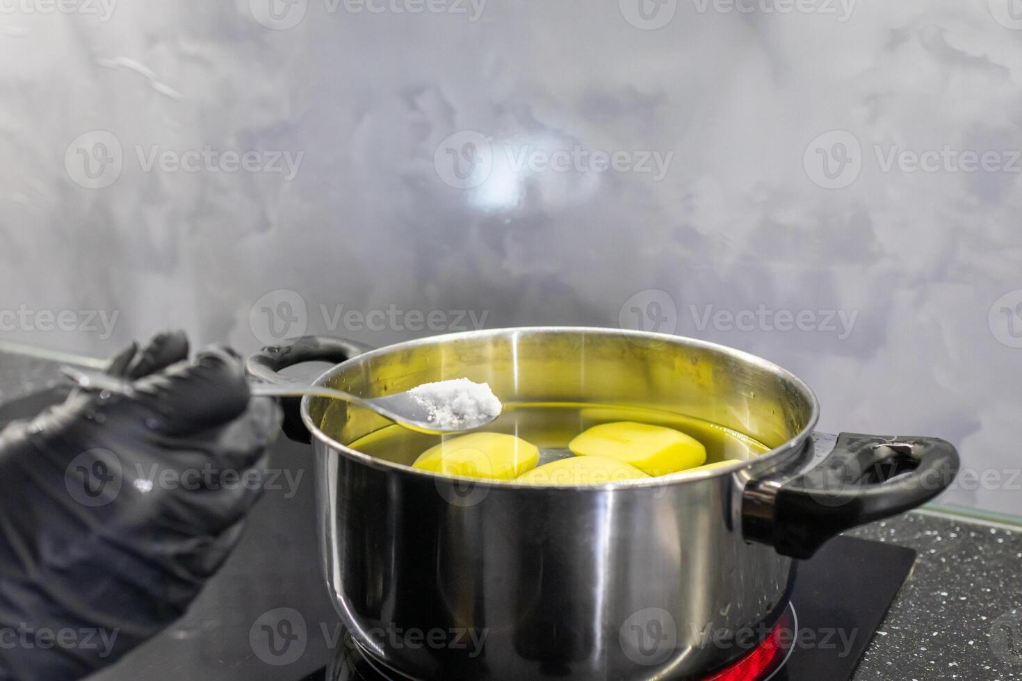 Person in black glove adding salt to a pot of boiling potatoes on a stove, symbolizing home cooking and healthy meal preparation photo