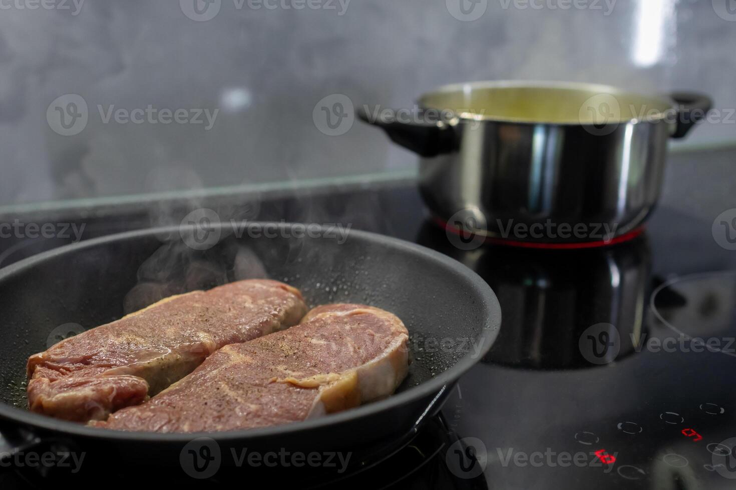 Two raw meat steaks sizzling in a hot frying pan on a modern induction hob, showcasing delicious home cooking, meal preparation, and culinary activity photo