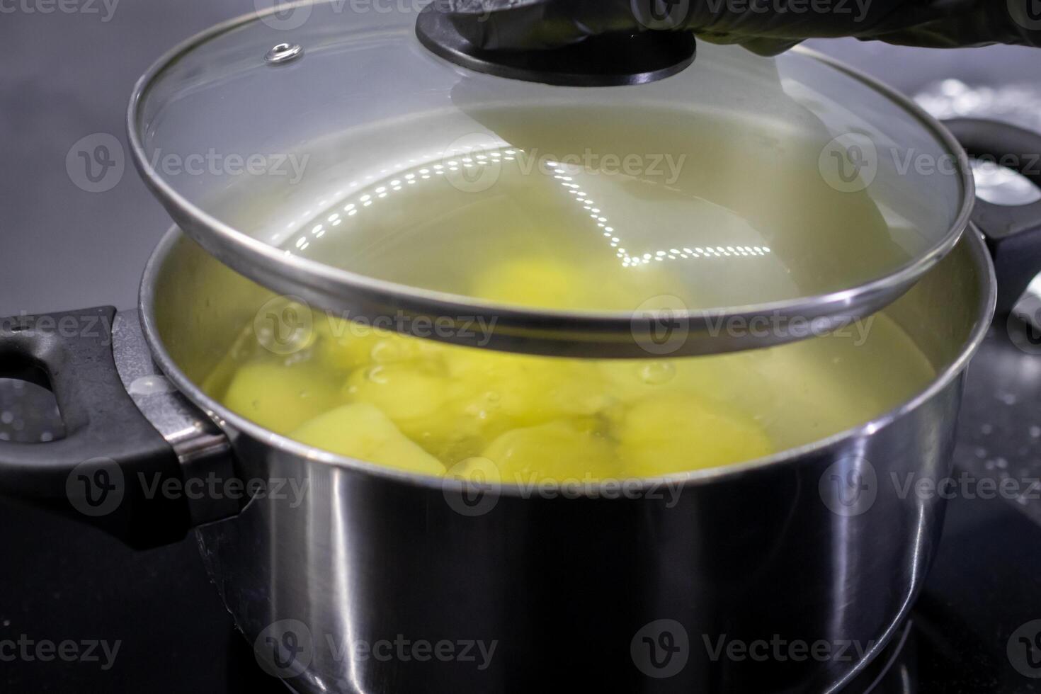A black-gloved hand lifts the glass lid from a stainless steel pot of boiling potatoes, illustrating healthy home cooking and everyday meal preparation photo