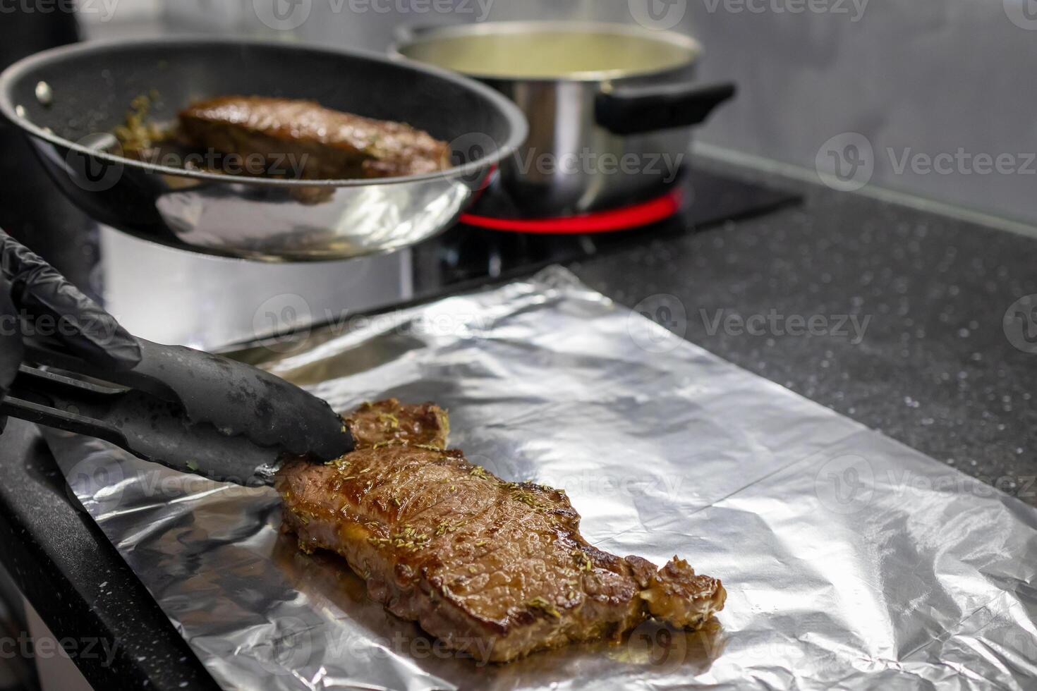 Skilled gloved hand transferring a perfectly seared steak from a hot frying pan to aluminum foil using tongs, symbolizing gourmet home cooking photo